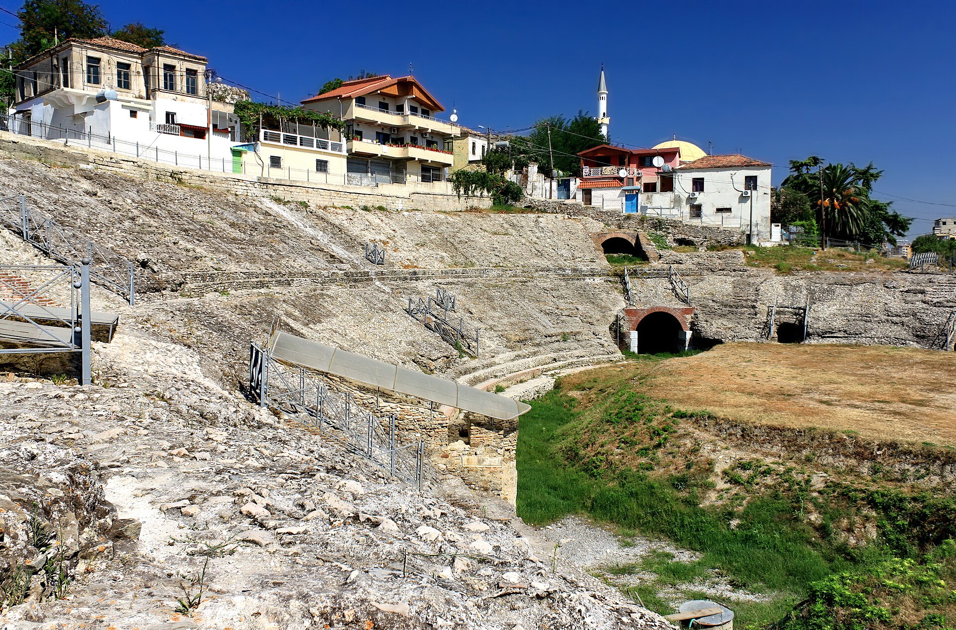 Ancient Roman amphitheater of Durres with stone seating and houses on its rim under a blue sky